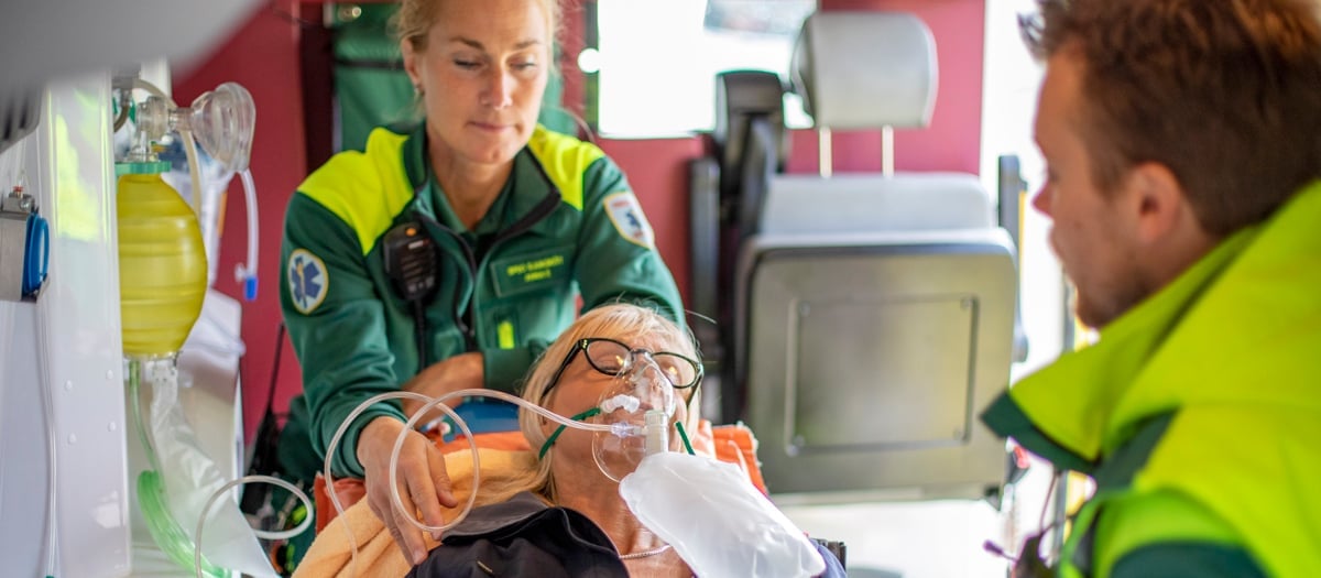 Två medarbetare inom ambulanssjukvården i neongröna arbetskläder tar hand om en kvinnlig patient som sitter på en bår.