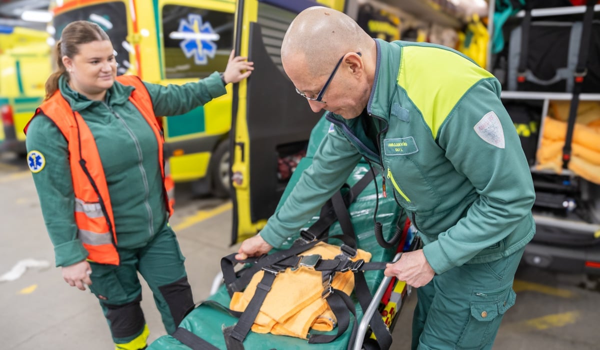 Två personer i ambulanskläder jobbar med en bår utanför ambulansen.