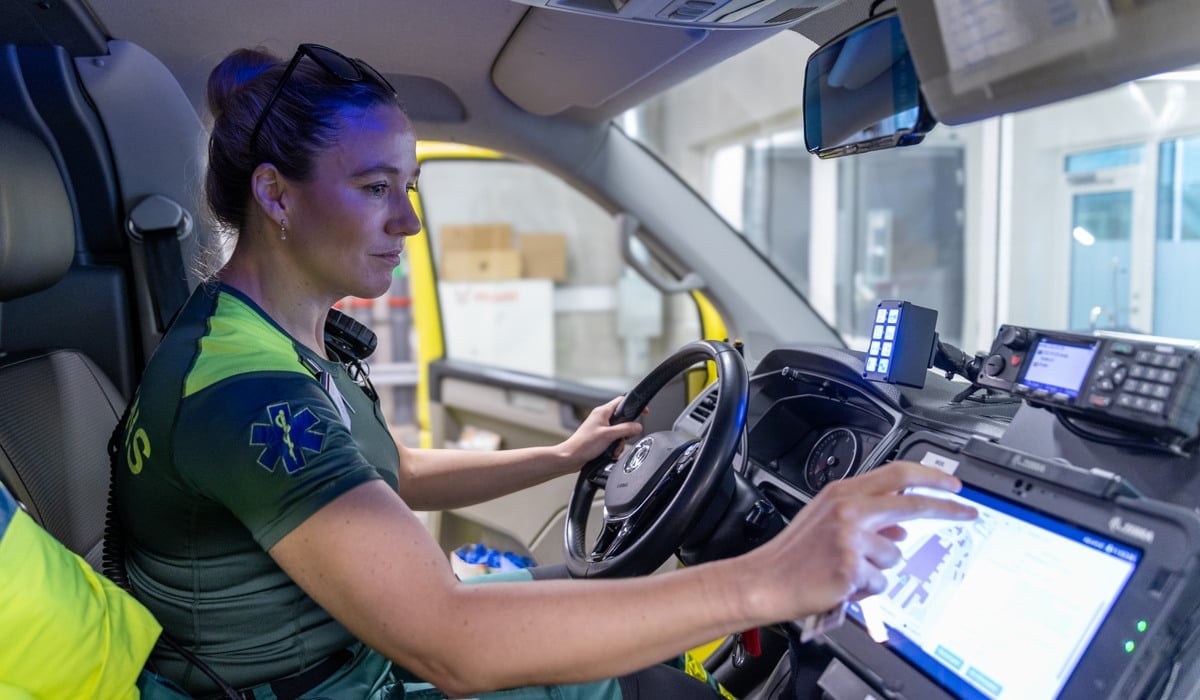 Kvinna i ambulanskläder sitter i förarhytt i ambulans och trycker på en skärm