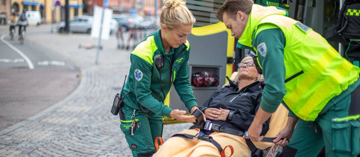 Två medarbetare inom ambulanssjukvården i neongröna arbetskläder tar hand om en kvinnlig patient som sitter på en bår.