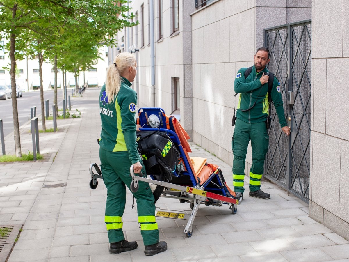 Två medarbetare i gröna arbetskläder väntar på att bli insläppa genom en grind. Den kvinnliga medarbetaren håller i en bår