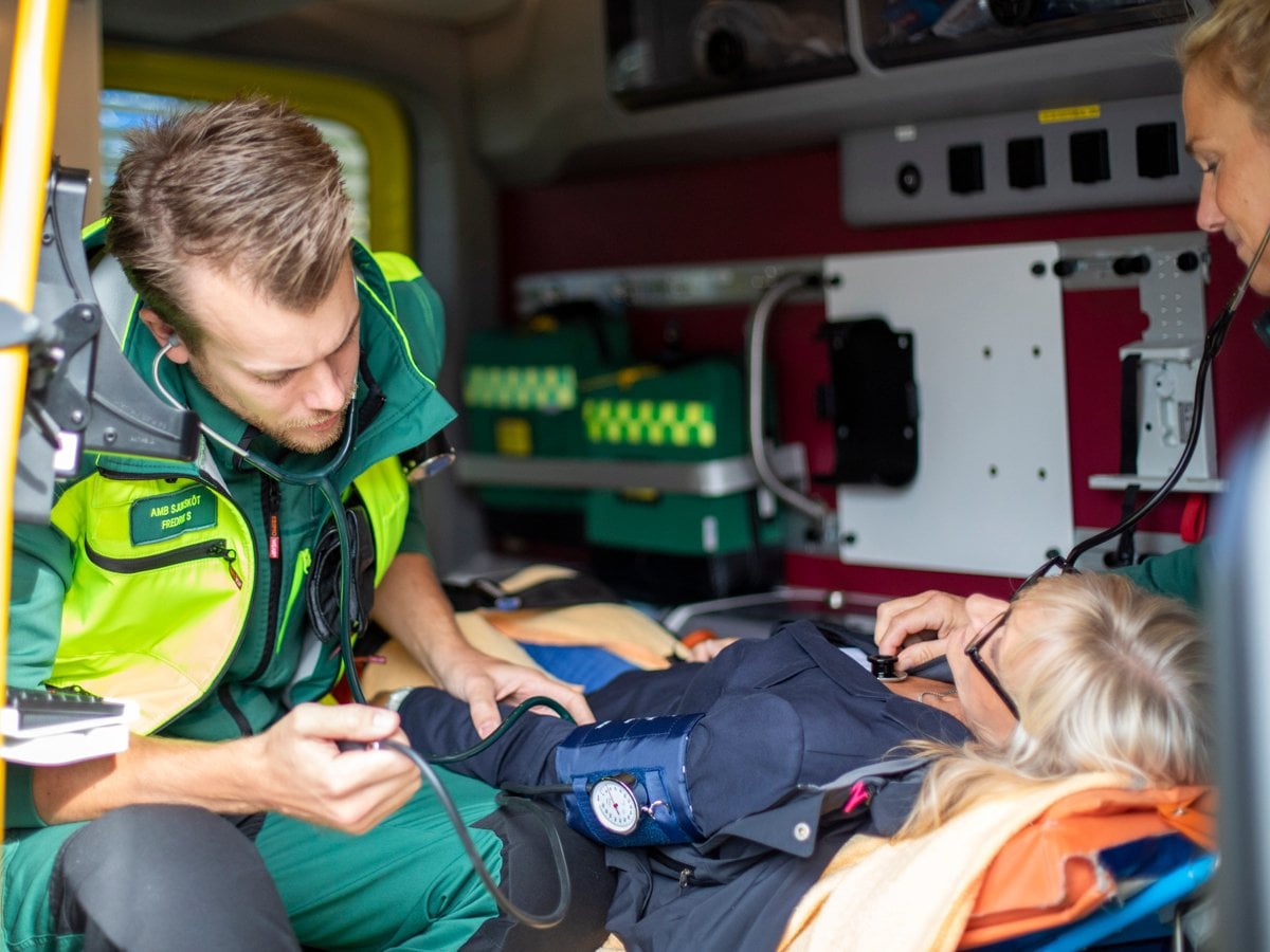 Två medarbetare inom ambulanssjukvården i neongröna arbetskläder tar hand om en kvinnlig patient som sitter på en bår.